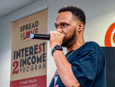 Man speaking at detention center graduation with Spread the Love, Interest 2 Income program poster in the background..